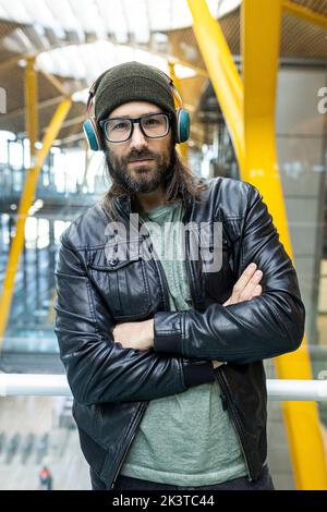 Handsome bearded hipster guy in eyeglasses and headphones wearing black leather jacket and knitted hat looking at camera while standing in airport hal Stock Photo