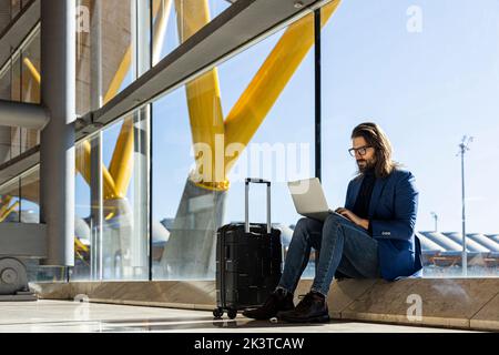 Serious adult businessman in trendy outfit and eyewear sitting next to glass wall and working with laptop while waiting for flight in airport lounge Stock Photo