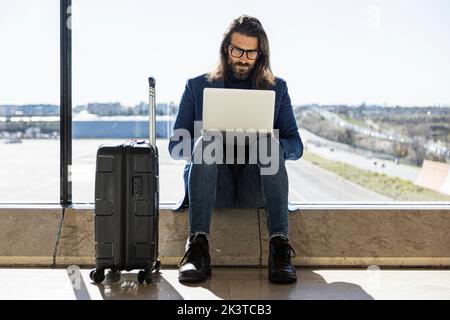 Serious adult businessman in trendy outfit and eyewear sitting next to glass wall and working with laptop while waiting for flight in airport lounge Stock Photo