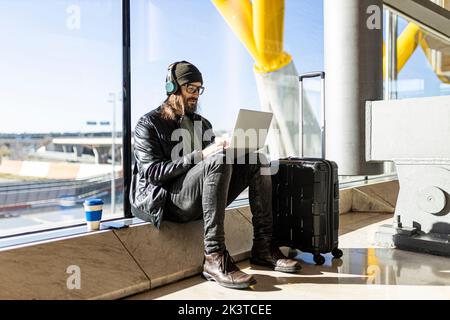 Positive adult hipster male traveler in black jacket and jeans using wireless headphones and laptop while waiting for flight in airport lounge Stock Photo