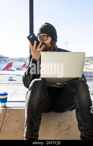 Stylish bearded guy in knitted hat and eyeglasses sending using phone while sitting with laptop against glass wall in airport Stock Photo