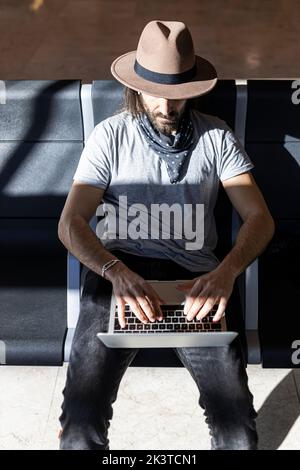 From above focused man with long hair in stylish outfit with hat sitting in a bench next to glass wall working on a laptop while waiting for flight in Stock Photo