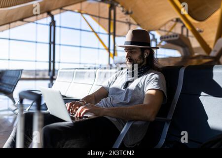 Side view of focused man with long hair in stylish outfit with hat sitting in a bench next to glass wall working on a laptop while waiting for flight Stock Photo