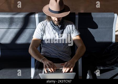 From above focused man with long hair in stylish outfit with hat sitting in a bench next to glass wall working on a laptop while waiting for flight in Stock Photo
