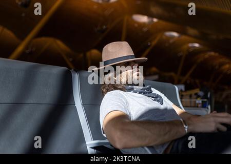 Adult bearded man in stylish outfit with hat sitting in a bench sleeping next to glass wall listening to music on wireless earphones while waiting for Stock Photo