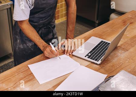 Cropped unrecognizable male bakery owner standing at counter writing notes on paper and using laptop and ingredients for baking working Stock Photo