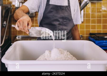 Crop anonymous male baker putting flour into plastic container while preparing dough for pastry in bakeshop kitchen Stock Photo