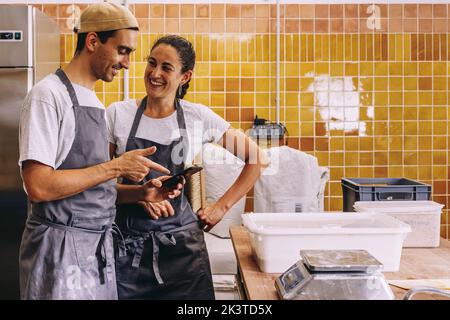 Happy young couple of small business owners using mobile phone and making order to supplier while working together in bakery Stock Photo
