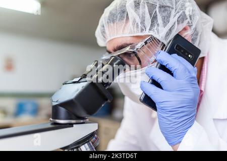 Man working at a lab using a smartphone while using a microscope at a laboratory Stock Photo