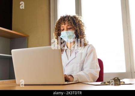 Ethnic female medic in disposable mask and uniform typing on netbook at table in clinic Stock Photo