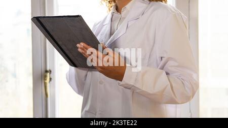 Anonymous crop content ethnic female doctor in medical uniform with tablet against window in hospital Stock Photo
