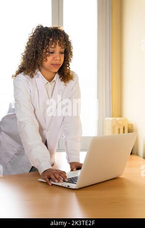 Concentrated ethnic female medic in uniform with hand in the table against netbook in clinic Stock Photo