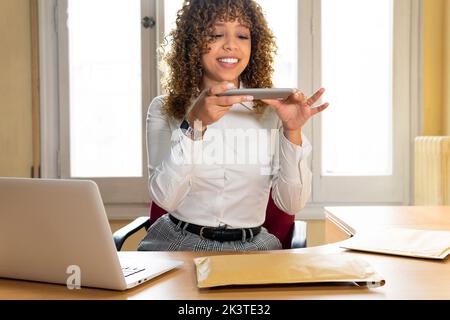 Crop content ethnic female entrepreneur taking photo of envelope on cellphone at table with laptop in office Stock Photo