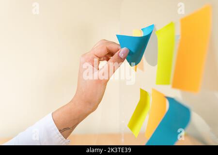 Anonymous ethnic female worker putting colorful sticky notes on wall in office Stock Photo
