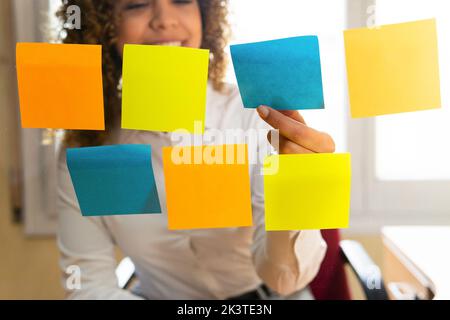 Through glass view of crop content ethnic female worker putting colorful sticky notes on wall in office Stock Photo