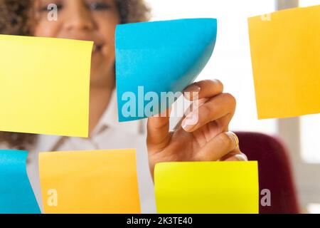 Through glass view of crop content ethnic female worker putting colorful sticky notes on wall in office Stock Photo