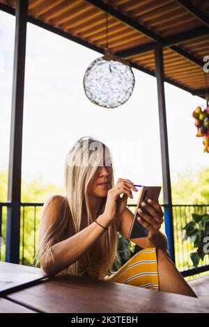 Young female in casual summer wear using mobile phone and netbook while sitting on bench on veranda in lush garden Stock Photo