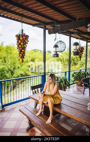 Full body young fit female in yellow sundress sitting on wooden table and looking away while sitting on resort terrace in lush summer garden Stock Photo