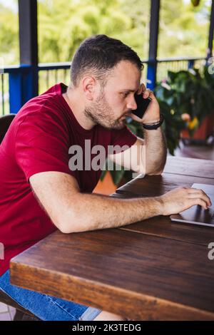 Concentrated young male in casual clothes talking mobile phone and browsing netbook while sitting at table on outdoor restaurant terrace Stock Photo