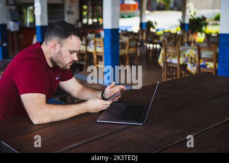 Concentrated young male in casual clothes browsing mobile phone and netbook while sitting at table on outdoor restaurant terrace Stock Photo