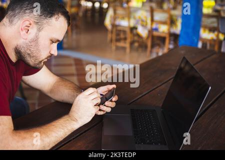 Concentrated young male in casual clothes browsing mobile phone and netbook while sitting at table on outdoor restaurant terrace Stock Photo