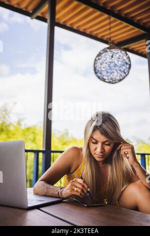 Young female in casual summer wear using mobile phone and netbook while sitting on bench on veranda in lush garden Stock Photo