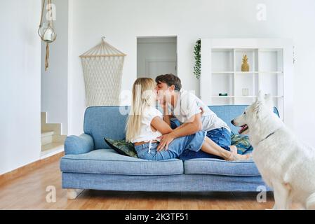 Side view of couple in love sitting on couch in room with White Swiss Shepherd dog and tenderly kissing Stock Photo