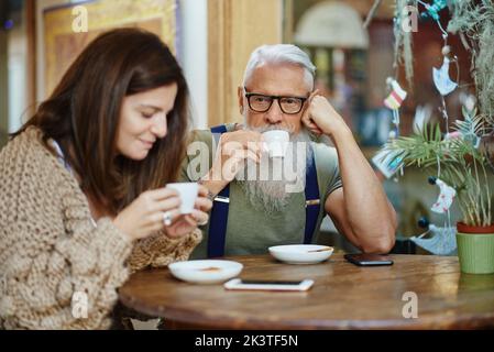 Adult smiling woman spending time with bearded stylish grandad while drinking from cups with hot drinks in cafeteria Stock Photo