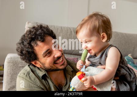 Delighted young ethnic man with dark hair smiling while playing with adorable little daughter siting on floor near sofa at home Stock Photo
