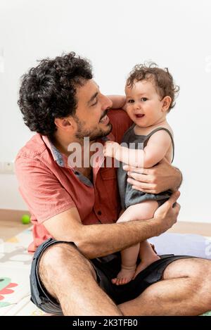 Excited young ethnic father with curly dark hair in casual clothes sitting on soft mat and playing with adorable little baby at home Stock Photo