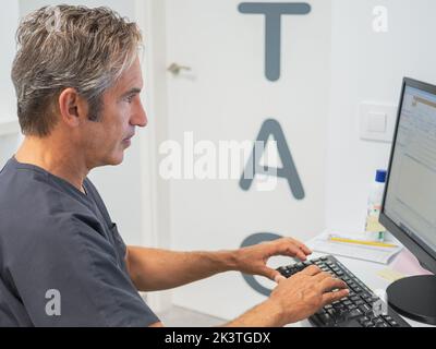 Side view of mature man in scrubs typing on computer keyboard while working in lab of modern hospital Stock Photo
