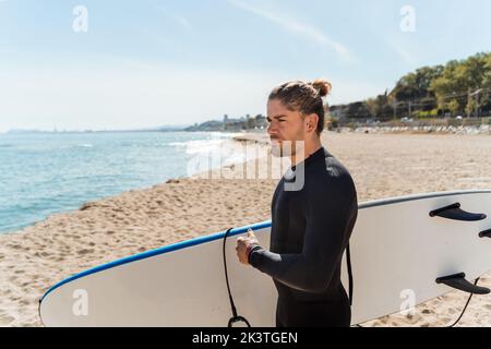 Side view of young thoughtful male with surfboard in hands standing on sandy beach near waving sea and looking away while preparing for surfing togeth Stock Photo