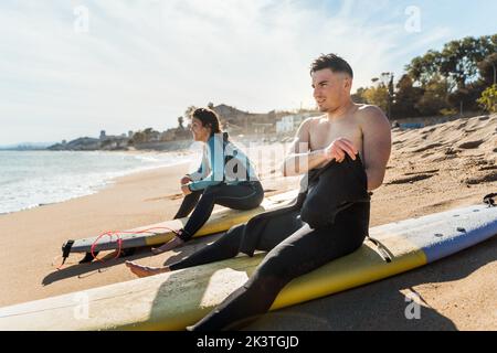Side view of young sporty man and woman in wetsuits sitting on surfboards on sandy beach while resting after surfing workout in summer day Stock Photo