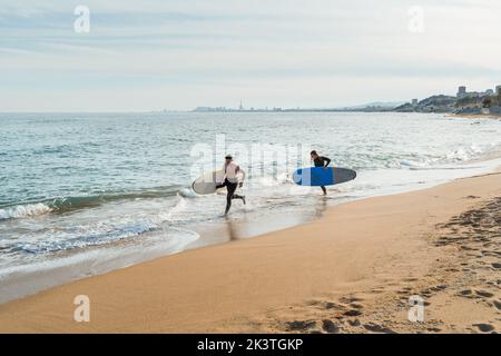 Young man and woman with surfboards in hands running along sandy seashore near waving sea while preparing for surfing together in summer day Stock Photo