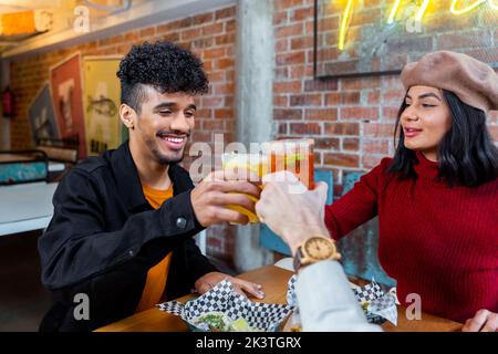 Excited young multiracial friends clinking glasses with tasty cocktails while gathering at table in modern restaurant Stock Photo