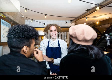 Smiling young waiter taking order and talking to ethnic couple while standing at counter in cafeteria Stock Photo