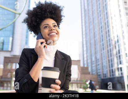 Smiling African American female architect in formal suit sitting on street with takeaway coffee and talking on smartphone while discussing project and Stock Photo