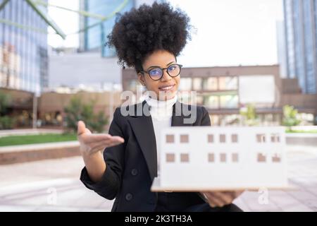 Content African American female architect in suit showing model of building made of paper and looking at camera while standing on city street Stock Photo