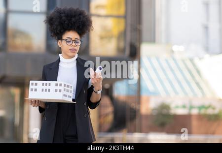 Serious African American female architect standing miniature model and sketch plan of house in city looking away Stock Photo