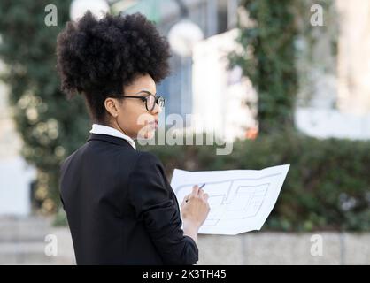Back view of determined African American female architect in suit standing on street with blueprint of building and looking away Stock Photo