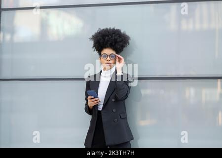 African American female architect in elegant outfit standing near glass wall browsing on mobile phone in city Stock Photo