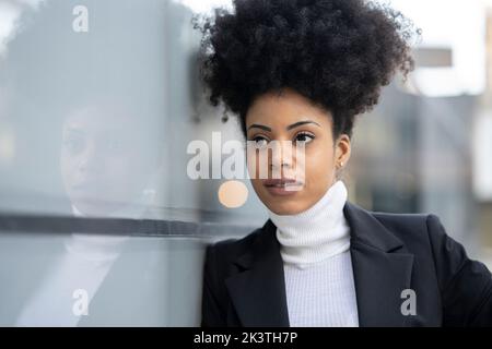 Black female architect in trendy suit and with Afro hairstyle leaning on glass wall of building while standing looking away Stock Photo