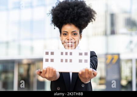 Content African American female architect in suit showing model of building made of paper and looking at camera while standing on city street Stock Photo
