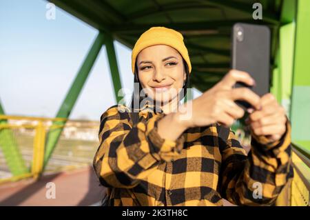 Positive young Latin American female with long dark hair in stylish checkered shirt and wool cap standing on bridge and taking selfie with mobile phon Stock Photo