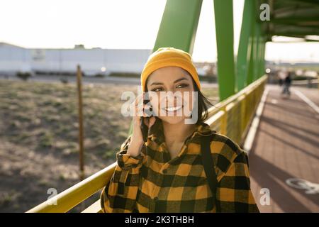 Positive young Latin American female with long dark hair in stylish checkered shirt standing on bridge and talking mobile phone on sunny day Stock Photo