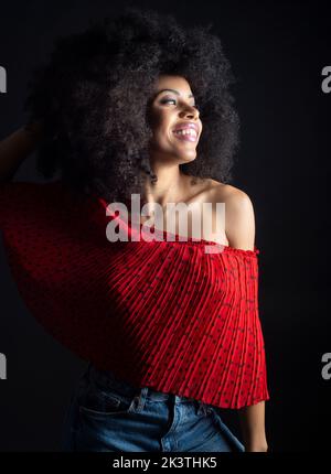 Cheerful young ethnic female with afro hair in ornamental red apparel with makeup and Afro hairstyle looking away in black studio background Stock Photo