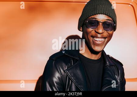 Handsome African American guy in stylish outfit smiling and looking at camera while standing near van on city street Stock Photo