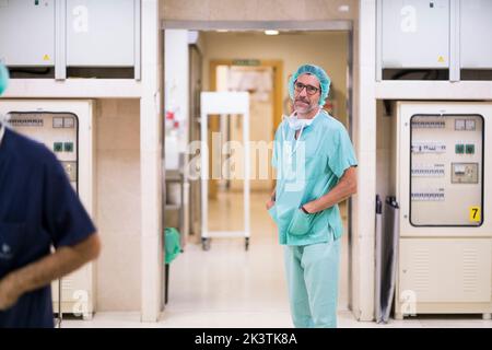 Side view of handsome adult man in surgeon uniform looking away and keeping hands in pockets while standing near entrance of operating theater Stock Photo