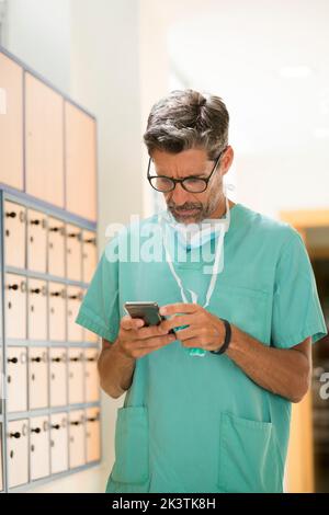 Adult man in surgeon uniform and glasses standing in hospital corridor and browsing modern smartphone Stock Photo