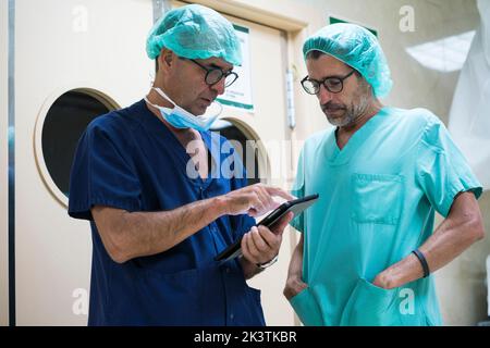 Two men in surgeon uniform standing in hospital corridor and using modern tablet together Stock Photo